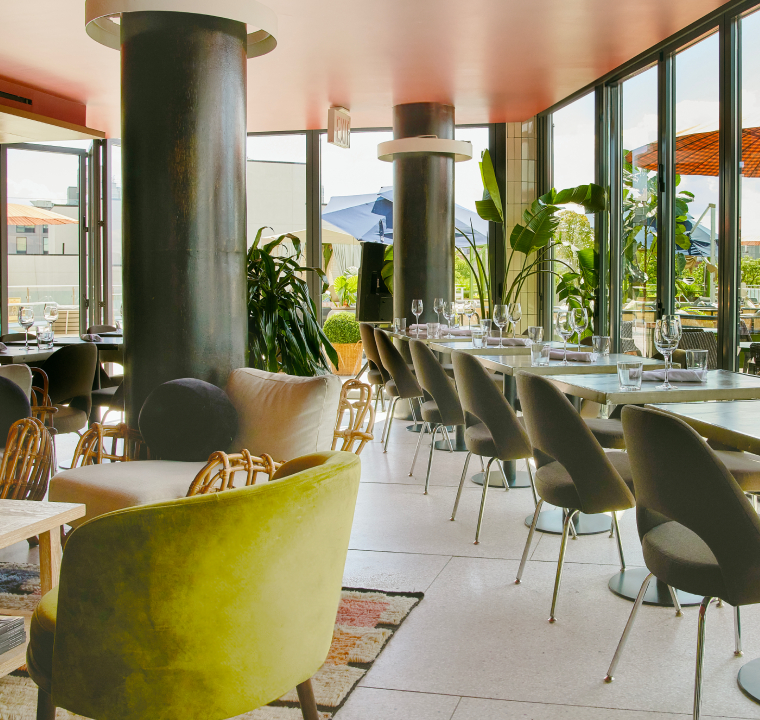 A long, rectangular dining table is set up outdoors on a rooftop, with a cityscape visible in the blurred background. The table is made of light-colored wood and is adorned with a lush green and white floral runner down its center. White plates are neatly stacked at each place setting, accompanied by clear wine glasses. Numerous white chairs with light-colored upholstery are arranged along both sides of the table, indicating a large gathering. The overall impression is one of an elegant outdoor dining event.
